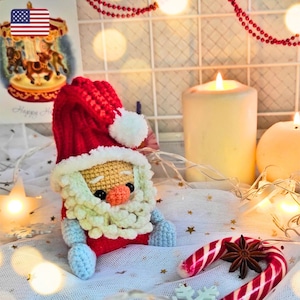 May include: A crocheted Santa Claus doll with a red hat, white beard, and blue mittens. The doll is sitting on a white fabric with a candy cane and a star anise.