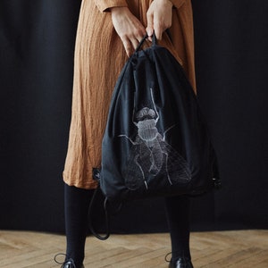 Könnte beinhalten: Eine schwarze Kordeltasche mit einem weißen gestickten Fliegenmotiv. Die Tasche wird von einer Person gehalten, die ein braunes Kleid und schwarze Schuhe trägt.