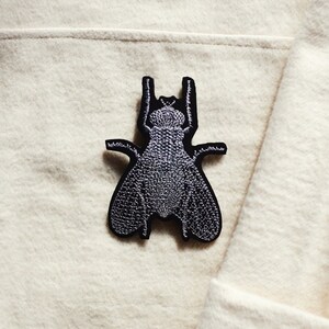 Peut inclure: Un patch thermocollant brodé noir représentant une mouche. La mouche est détaillée avec des points de couture et est de couleur grise.