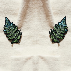 May include: Two green and blue embroidered fern leaf patches on a white fabric background.