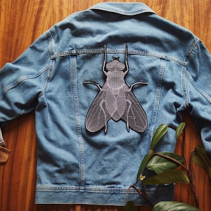 Peut inclure: Une veste en jean bleue avec un patch brodé de mouche gris sur le dos.