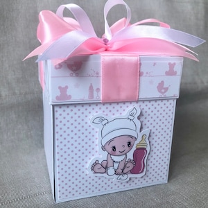 Könnte beinhalten: Eine weiße Geschenkbox mit rosafarbenem Punkte-Muster und einem rosafarbenen Band. Die Box hat ein Bild eines kleinen Mädchens mit einer Flasche auf der Vorderseite.