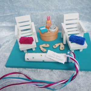 Könnte beinhalten: Eine Miniatur-Strandkulisse mit zwei weißen Liegestühlen, einem kleinen Tisch mit einer Flasche Sonnencreme, einem Buch und einem Tiegel Lotion. Auf der blauen Fläche liegen Flip-Flops und eine weiße Tube mit einem Zettel, auf dem steht "Für einen kleinen Ausflug!"