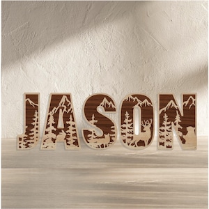 May include: Wooden name sign spelling "JASON". Each letter is a combination of dark and light wood, with mountain and forest scenes cut out within the letters. The letters are standing on a wooden surface.