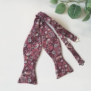 Puede incluir: Una pajarita burdeos con un estampado floral y de pájaros. La pajarita ajustable presenta un diseño repetido de flores, pájaros y hojas en tonos rosa, blanco y burdeos. La pajarita es de tela y tiene un cierre metálico.