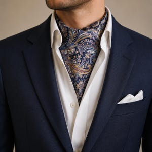 Puede incluir: Una americana azul marino sobre una camisa blanca de botones con un pañuelo de bolsillo blanco. Un ascot con un detallado estampado de cachemira en tonos azul marino, dorado y azul claro está metido en el cuello de la camisa blanca.