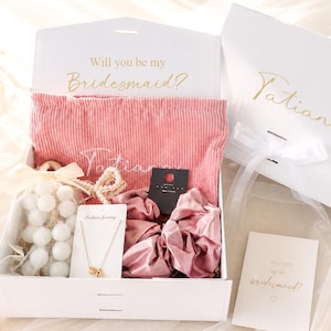 May include: A white gift box with the text "Will you be my Bridesmaid?" and "Tatiana" in gold script. Inside, a pink corduroy pouch, jewelry, scrunchies, and a card. A second box is visible.