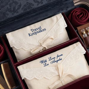 Puede incluir: Un maletín de viaje con forro oscuro que contiene dos bolsas de tela color crema con ribete de encaje y texto bordado en azul Travel Keepsakes y With Love from Los Angeles, dos peines marrones, dos rollos de tela burdeos y viales de vidrio.