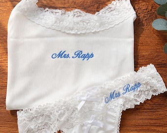 May include: White lingerie set with a camisole and thong, both trimmed with lace. The camisole and thong feature the embroidered text "Mrs. Rapp" in blue. A pair of white heart-shaped sunglasses sits above the lingerie.