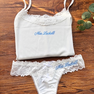 Può includere: Completo intimo bianco composto da canotta a costine e perizoma abbinato, con bordi in pizzo e testo ricamato blu "Mrs. Luckett". Occhiali da sole bianchi a forma di cuore in alto.