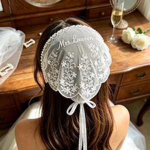 Peut inclure: Une coiffe en dentelle blanche avec un bord festonné et une broderie florale complexe. Le texte Mrs. Lauren est brodé en écriture blanche sur le dessus. L'accessoire comprend un ruban blanc délicat à la base pour le maintenir en place.
