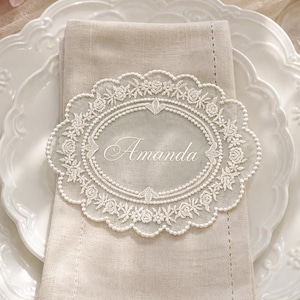 May include: A beige linen napkin folded on a white scalloped plate, topped with an oval white lace doily featuring intricate floral embroidery and the name Amanda stitched in the centre, accompanied by a silver fork and knife with cream handles.
