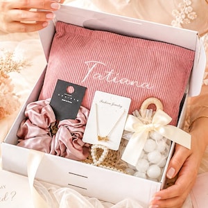 May include: A white gift box filled with a pink pillow embroidered with "Tatiana", scrunchies, jewelry, and a small bag of white candies. The box also contains cards that read "Will you be my Bridesmaid?" and "Thank you for being part of my special day."