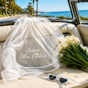 May include: A white tulle wedding veil featuring the embroidered text Future Mrs. Carlson, accompanied by a bouquet of white tulips and white heart-shaped sunglasses resting on a cream-colored car seat with a coastal road view in the background.