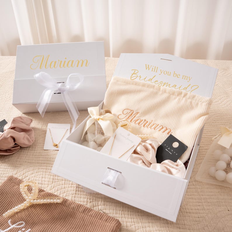 May include: A white gift box with a ribbon, containing a beige fabric, jewelry, and scrunchies. The box is labeled "Mariam" in gold script, with the text "Will you be my Bridesmaid?" on the inside lid. A gift set for a bridesmaid.