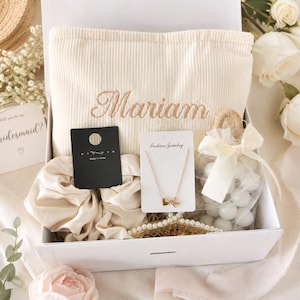 May include: A white gift box filled with a cream-colored tote bag embroidered with the name "Mariam", a gold bow necklace, a pearl bracelet, a scrunchie, and a small bag of white candies. A card that says "Will you be my bridesmaid?" is visible.