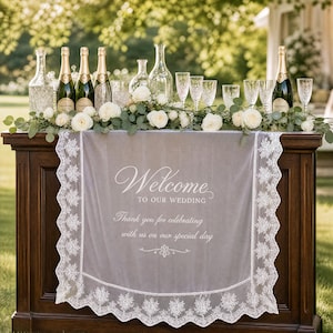 May include: A white lace table runner draped over a dark wooden table, featuring the printed text Welcome TO OUR WEDDING Thank you for celebrating with us on our special day. The table is decorated with white roses, greenery, glass bottles, and glasses.
