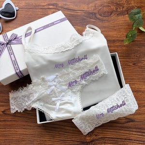 Pu&ograve; includere: Un completo intimo bianco con rifiniture in pizzo e fiocchi, presentato in una confezione regalo aperta. Il set include una canotta, slip e giarrettiera, tutti ricamati con "Mrs Mitchell" in viola. La scatola &egrave; bianca con un nastro "Best Wishes".