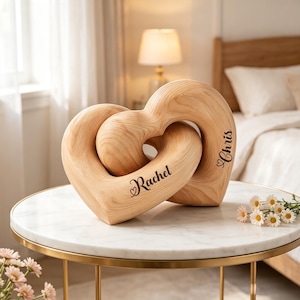 Puede incluir: Una escultura de madera de color claro que consiste en dos formas de corazón entrelazadas. La madera presenta un patrón de veta natural visible. El nombre Rachel está grabado en el corazón izquierdo y Chris en el derecho.