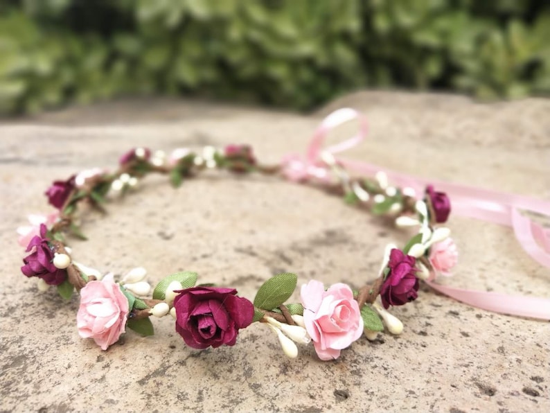 Puede incluir: Una corona floral rosa y burdeos con cuentas blancas y hojas verdes. La corona est&aacute; atada con una cinta rosa.