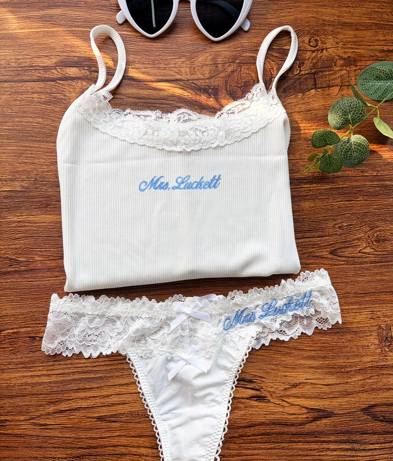 Pu&ograve; includere: Completo intimo bianco composto da canotta a costine e perizoma abbinato, con bordi in pizzo e testo ricamato blu "Mrs. Luckett". Occhiali da sole bianchi a forma di cuore in alto.