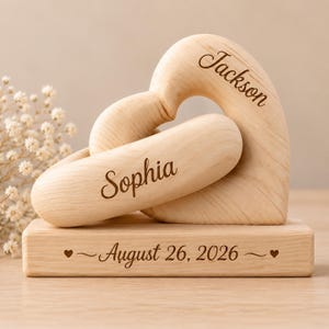 Puede incluir: Una escultura de madera de color claro con dos piezas entrelazadas en forma de corazón grabadas con los nombres Jackson y Sophia. La escultura descansa sobre una base rectangular de madera grabada con la fecha August 26, 2026, flanqueada por dos pequeños corazones.
