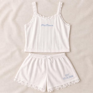 Puede incluir: Un conjunto de pijama blanco acanalado que consta de una camiseta de tirantes y pantalones cortos. La camiseta tiene ribetes de encaje, un lazo pequeño y el texto bordado en azul Mrs. Brown. El pantalón tiene un dobladillo de encaje y el texto EST. 12.12.2026.
