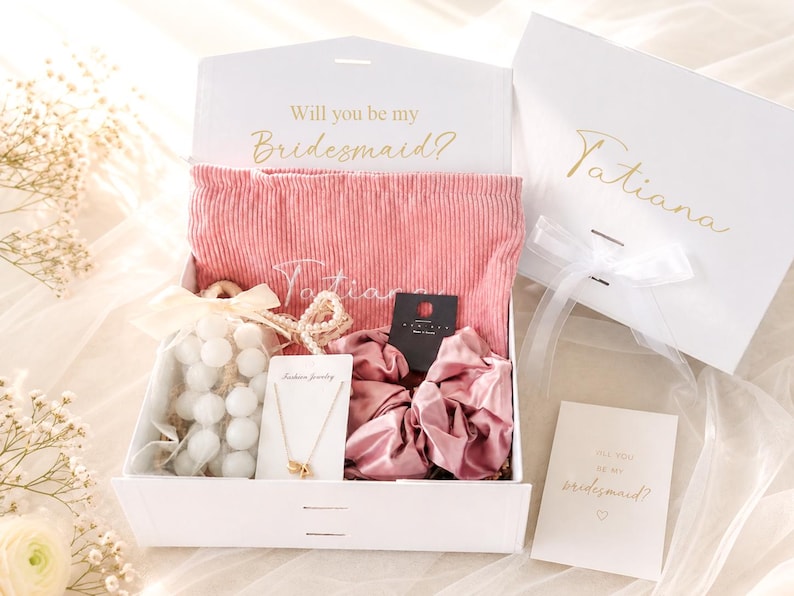 May include: A white gift box with the text "Will you be my Bridesmaid?" and "Tatiana" in gold script. Inside, a pink corduroy pouch, jewelry, scrunchies, and a card. A second box is visible.