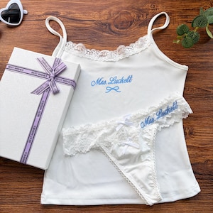 Pu&ograve; includere: Completo intimo bianco con rifiniture in pizzo e ricamo blu "Mrs. Luckett". Include una canotta e perizoma, insieme a una confezione regalo con un nastro "Best Wishes". Occhiali da sole a forma di cuore visibili.