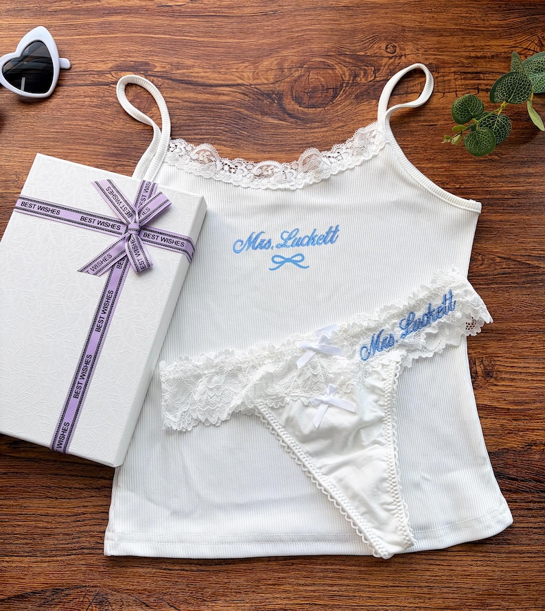 Pu&ograve; includere: Completo intimo bianco con rifiniture in pizzo e ricamo blu "Mrs. Luckett". Include una canotta e perizoma, insieme a una confezione regalo con un nastro "Best Wishes". Occhiali da sole a forma di cuore visibili.