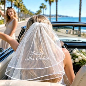 May include: A white two-tier bridal veil featuring a satin ribbon edge and white embroidered text that reads Future Mrs. Carlson. The veil is made of sheer tulle fabric and is worn over long blonde hair while sitting in a convertible car.