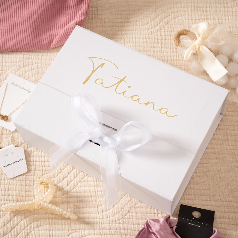 May include: A white gift box with the name "Tatiana" in gold script on the lid. The box is tied with a white ribbon. Jewelry and a hair clip are visible nearby, suggesting a gift set.