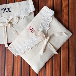 May include: Two off-white canvas pouches featuring white eyelet lace trim on the envelope-style flap. Each pouch is secured with a tied canvas ribbon. One pouch displays the embroidered navy blue letters VK, while the other features a red letter O.