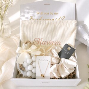 May include: A white gift box with a lid open, containing a cream-colored drawstring bag with the name "Mariam" embroidered in gold. The box also holds a necklace, scrunchies, and a bag of white spheres. The text "Will you be my Bridesmaid?" is printed on the inside of the lid.