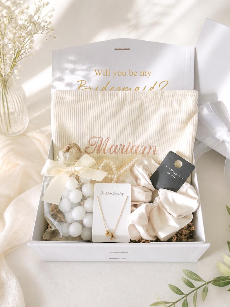 May include: A white gift box with a lid open, containing a cream-colored drawstring bag with the name "Mariam" embroidered in gold. The box also holds a necklace, scrunchies, and a bag of white spheres. The text "Will you be my Bridesmaid?" is printed on the inside of the lid.