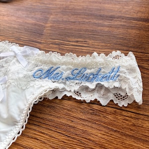 Pu&ograve; includere: Mutandine in pizzo bianco con testo ricamato blu "Mrs. Luckett". La biancheria intima presenta delicate finiture in pizzo e piccoli fiocchi bianchi. Il capo &egrave; esposto su una superficie di legno.