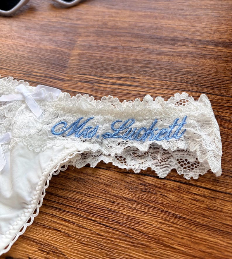 Pu&ograve; includere: Mutandine in pizzo bianco con testo ricamato blu "Mrs. Luckett". La biancheria intima presenta delicate finiture in pizzo e piccoli fiocchi bianchi. Il capo &egrave; esposto su una superficie di legno.
