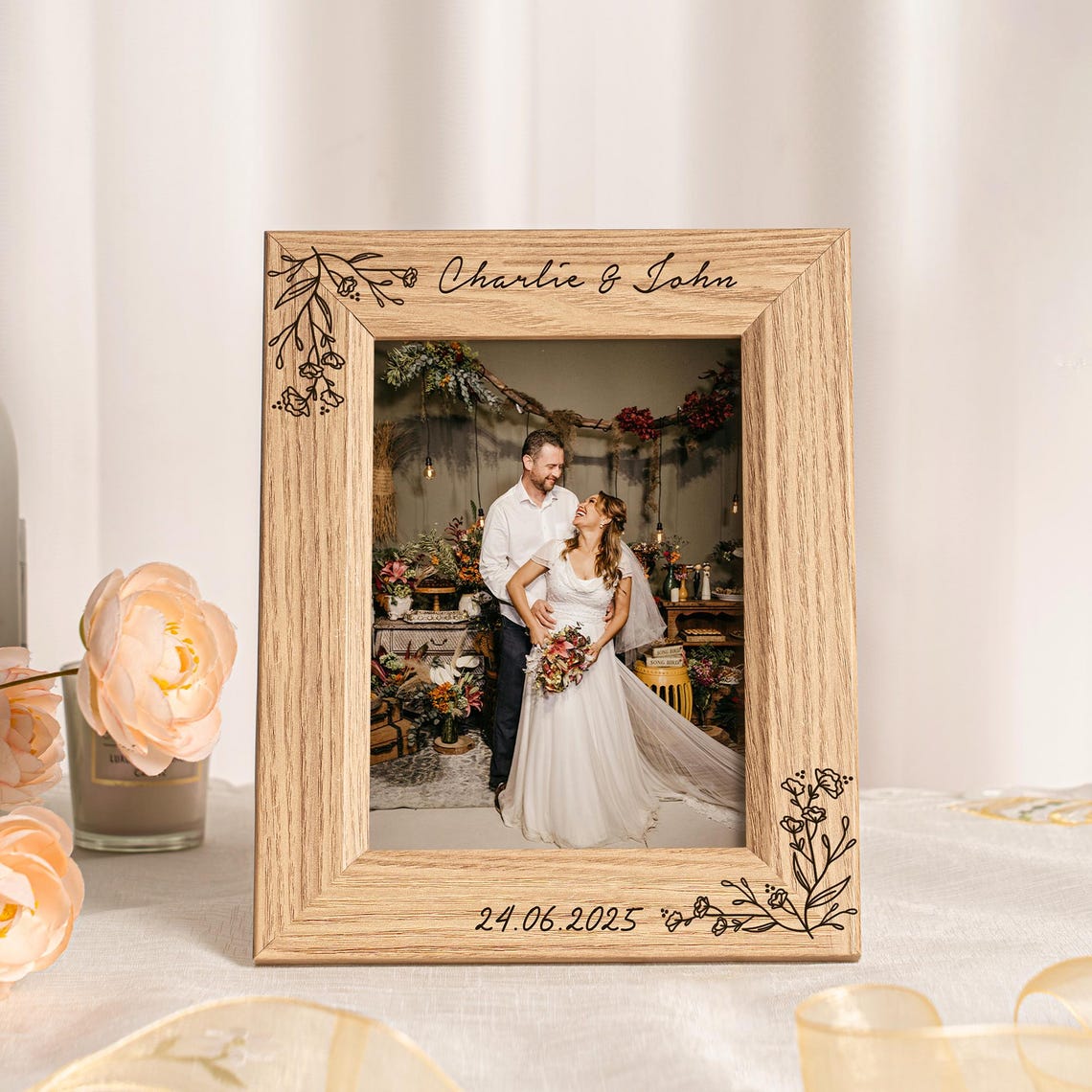 May include: A wooden picture frame with the names "Charlie & John" engraved at the top and the date "24.06.2025" at the bottom. The frame has floral designs in the corners and holds a wedding photo of a couple.