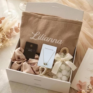 May include: A white gift box filled with a beige corduroy pouch embroidered with the name "Lilianna", a satin scrunchie, a necklace, a bag of white candies, and a card. The box is open, revealing the contents.