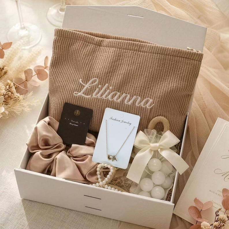 May include: A white gift box filled with a beige corduroy pouch embroidered with the name "Lilianna", a satin scrunchie, a necklace, a bag of white candies, and a card. The box is open, revealing the contents.