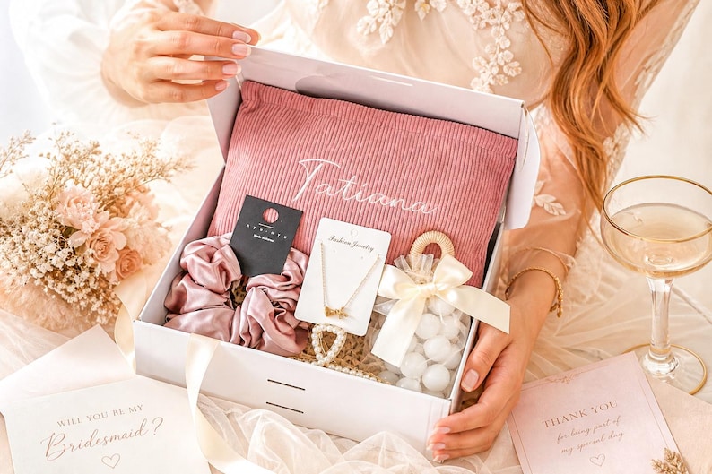 May include: A white gift box filled with a pink pillow embroidered with "Tatiana", scrunchies, jewelry, and a small bag of white candies. The box also contains cards that read "Will you be my Bridesmaid?" and "Thank you for being part of my special day."