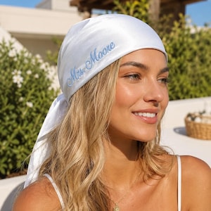 May include: A white satin headscarf featuring the embroidered text Mrs. Moore in light blue script lettering. The headscarf is tied at the back and has a smooth, shiny finish, covering the top of the head with a triangular shape and side tails.