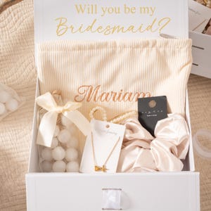May include: A white gift box with the text "Will you be my Bridesmaid?" in gold script. Inside, a cream-colored bag embroidered with a name, a gold necklace, a pearl bracelet, a scrunchie, and a bottle of white candies.