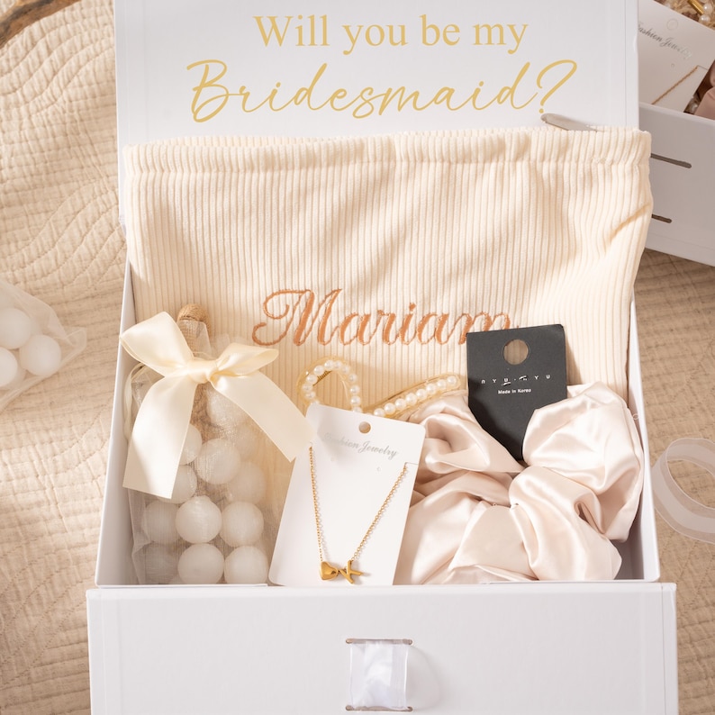 May include: A white gift box with the text "Will you be my Bridesmaid?" in gold script. Inside, a cream-colored bag embroidered with a name, a gold necklace, a pearl bracelet, a scrunchie, and a bottle of white candies.