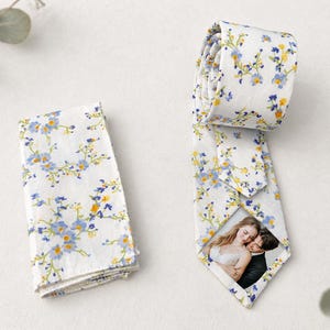 Puede incluir: Una corbata blanca y un pañuelo de bolsillo a juego con un estampado floral de pequeñas flores azules y amarillas. La parte inferior de la corbata muestra una fotografía rectangular de una pareja abrazada, integrada en el diseño.