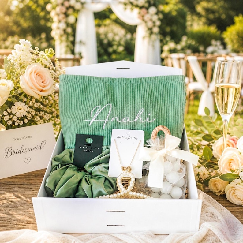 May include: A white gift box filled with a green corduroy pouch embroidered with a name, a necklace, a scrunchie, and a card that says "Will you be my Bridesmaid?" with a heart. A glass of champagne and flowers are in the background.