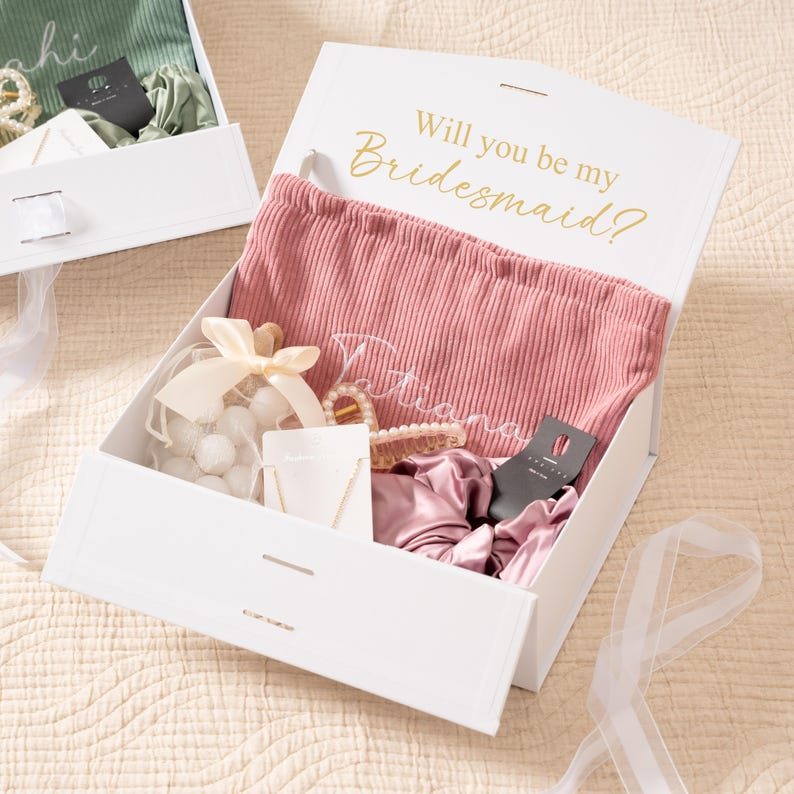 May include: White gift box with the text "Will you be my Bridesmaid?" in gold lettering. Inside, a pink corduroy pouch, jewelry, scrunchies, and a bag of white spheres are visible. A second box is partially visible.
