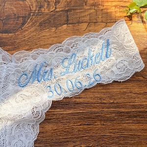 May include: White lace garter with blue embroidered text reading "Mrs. Luckett" and the date "30.06.26". The garter is displayed on a wooden surface, showcasing its delicate lace design and personalized details.