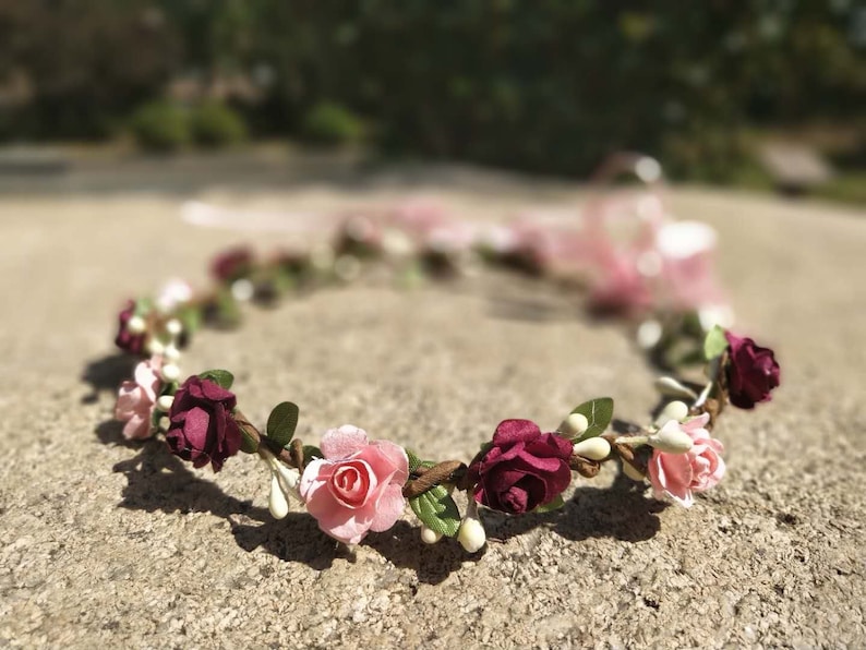 Puede incluir: Una corona floral rosa y burdeos con peque&ntilde;as flores blancas y hojas verdes.