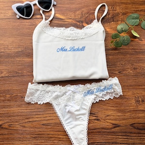 Pu&ograve; includere: Completo intimo bianco con top a camisole e perizoma. La camisole ha un bordo in pizzo e la scritta "Mrs. Luckett" in blu. Anche il perizoma presenta un bordo in pizzo e la stessa scritta. Occhiali da sole a forma di cuore.